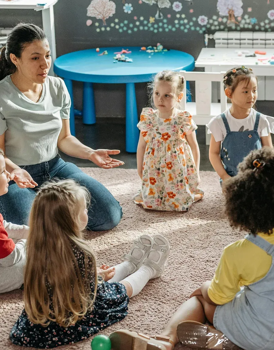 Un'educatrice seduta a terra in cerchio con un gruppo di bambini, in una stanza con un tavolo blu e disegni alle pareti.