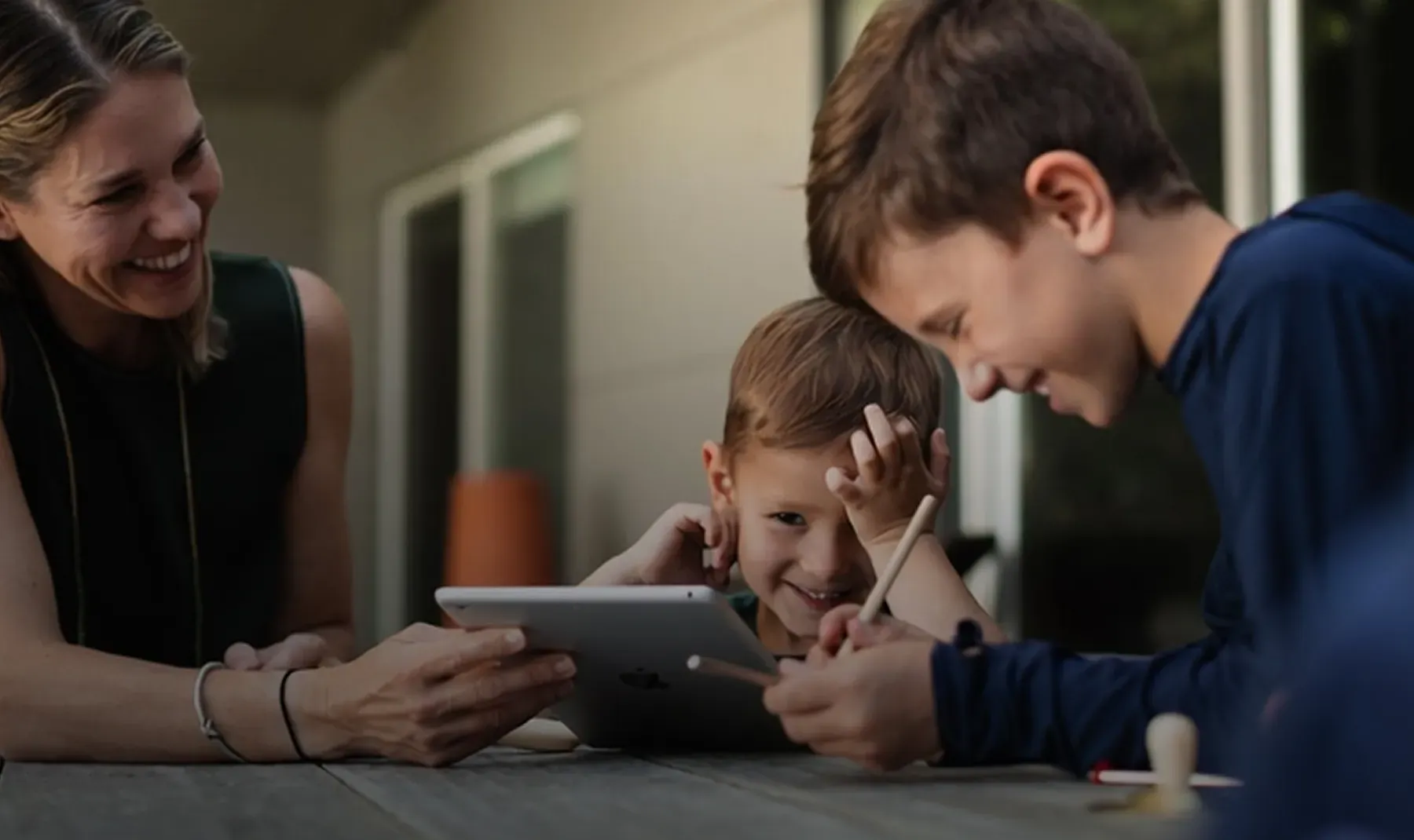 Una madre e due figli seduti a un tavolo all'aperto, sorridono guardando insieme un tablet.